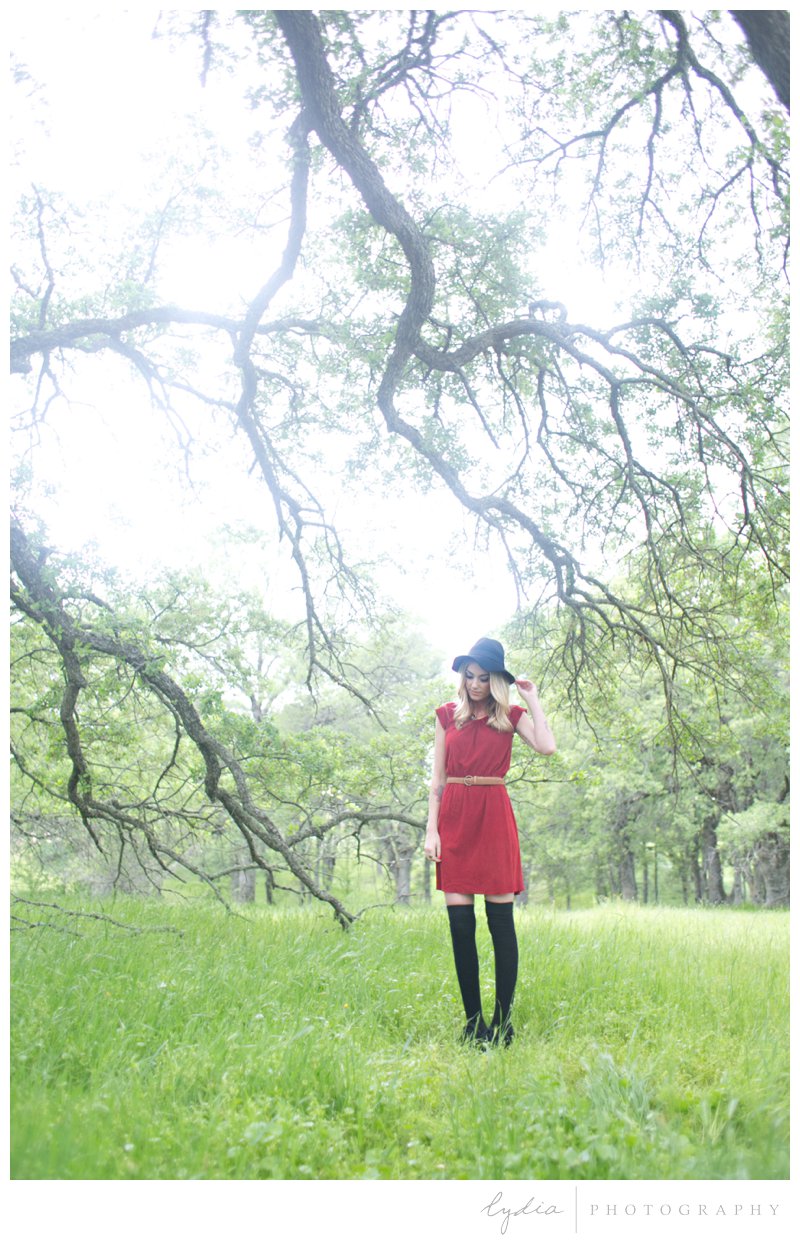 Anthropologie styled senior wearing a red vintage dress and black felt hat in a wildflower field under a tree in Roseville, California.