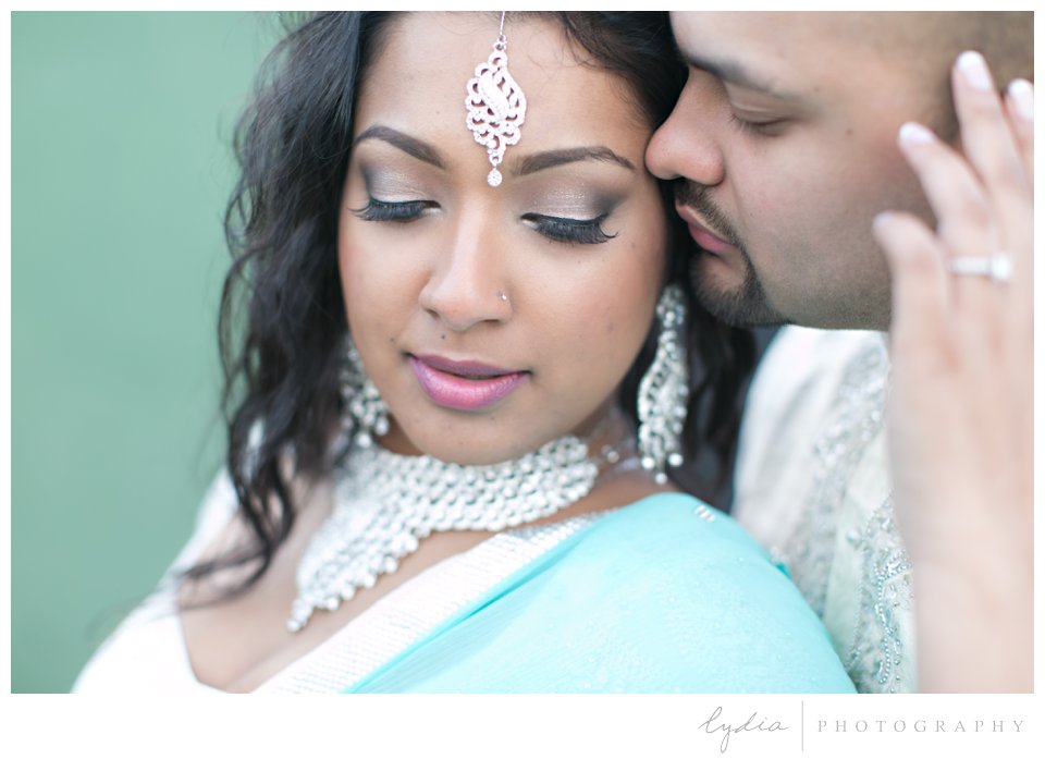 Indian bride wearing turquoise sari and silver bindi being kissed by groom