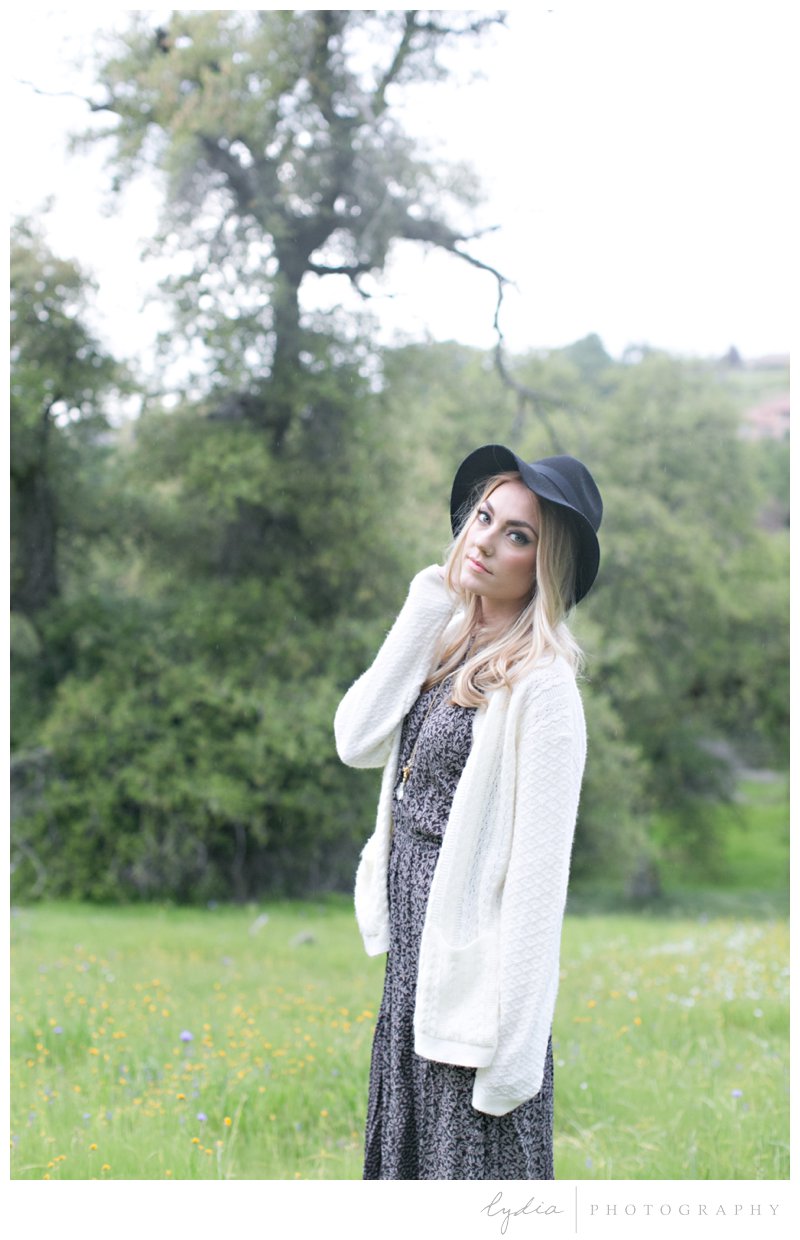 Anthropologie styled senior wearing a cozy sweater and black felt hat in a wildflower field in the rain in Roseville, California.