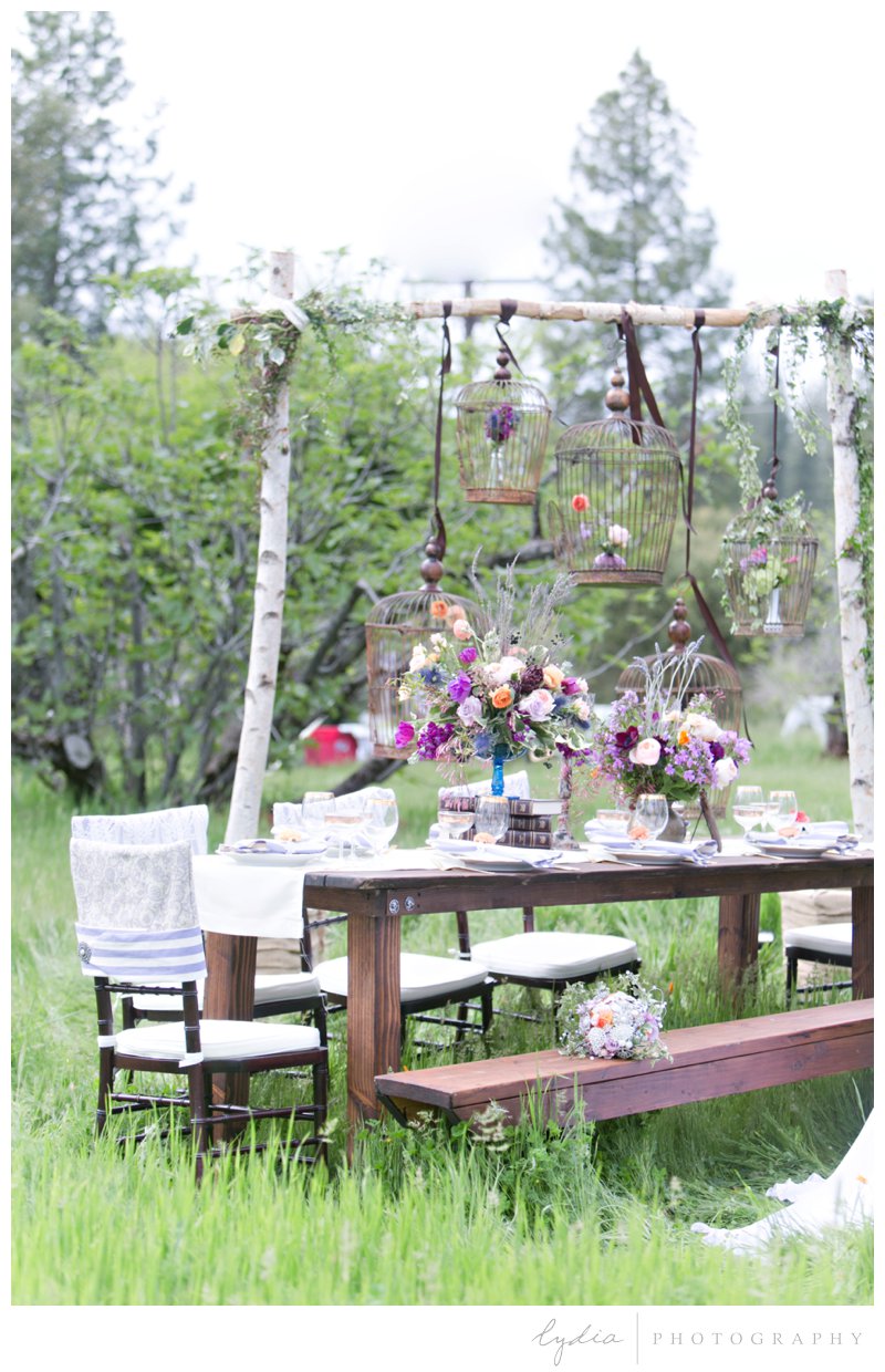 Birch frame with hanging floral birdcages and table setting with chiavari chairs, benches, antique books, and purple floral arrangements at French Countryside wedding at North Star House in Grass Valley, California.