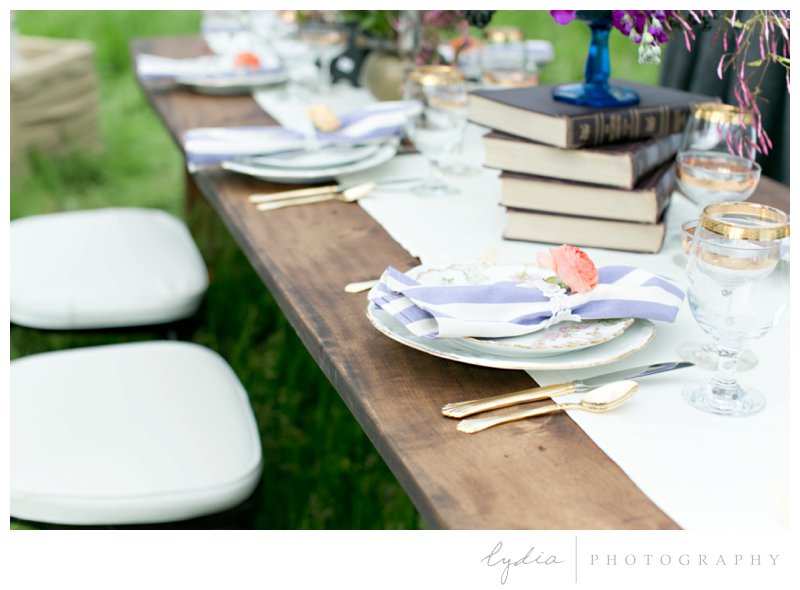 Table setting with gold rimmed glasses, gold silverware, antique china, and purple striped napkins at French Countryside wedding at North Star House in Grass Valley, California.